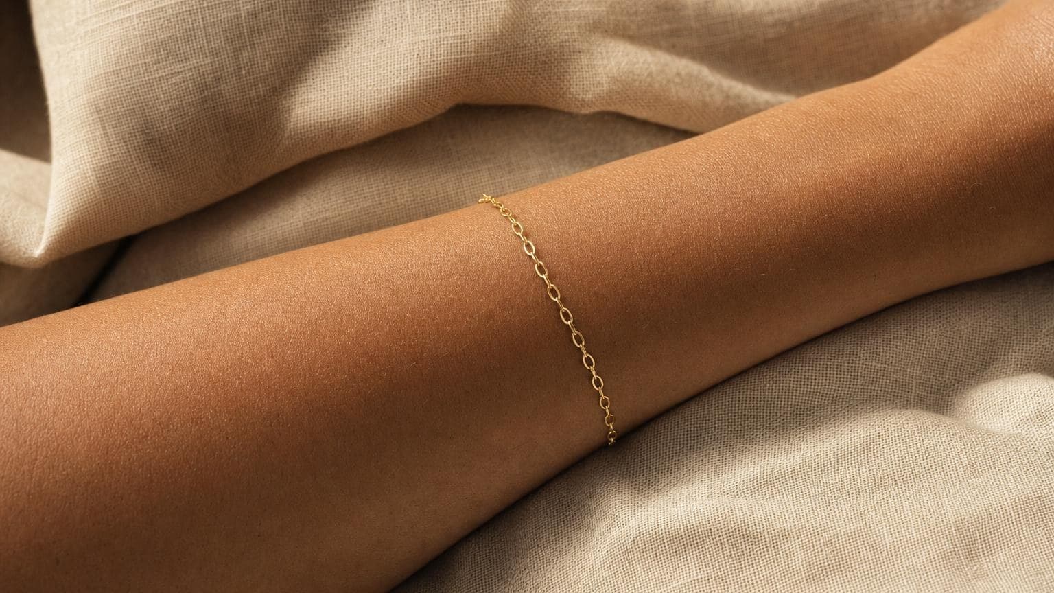 A single fine gold chain bracelet on a wrist resting on beige linen